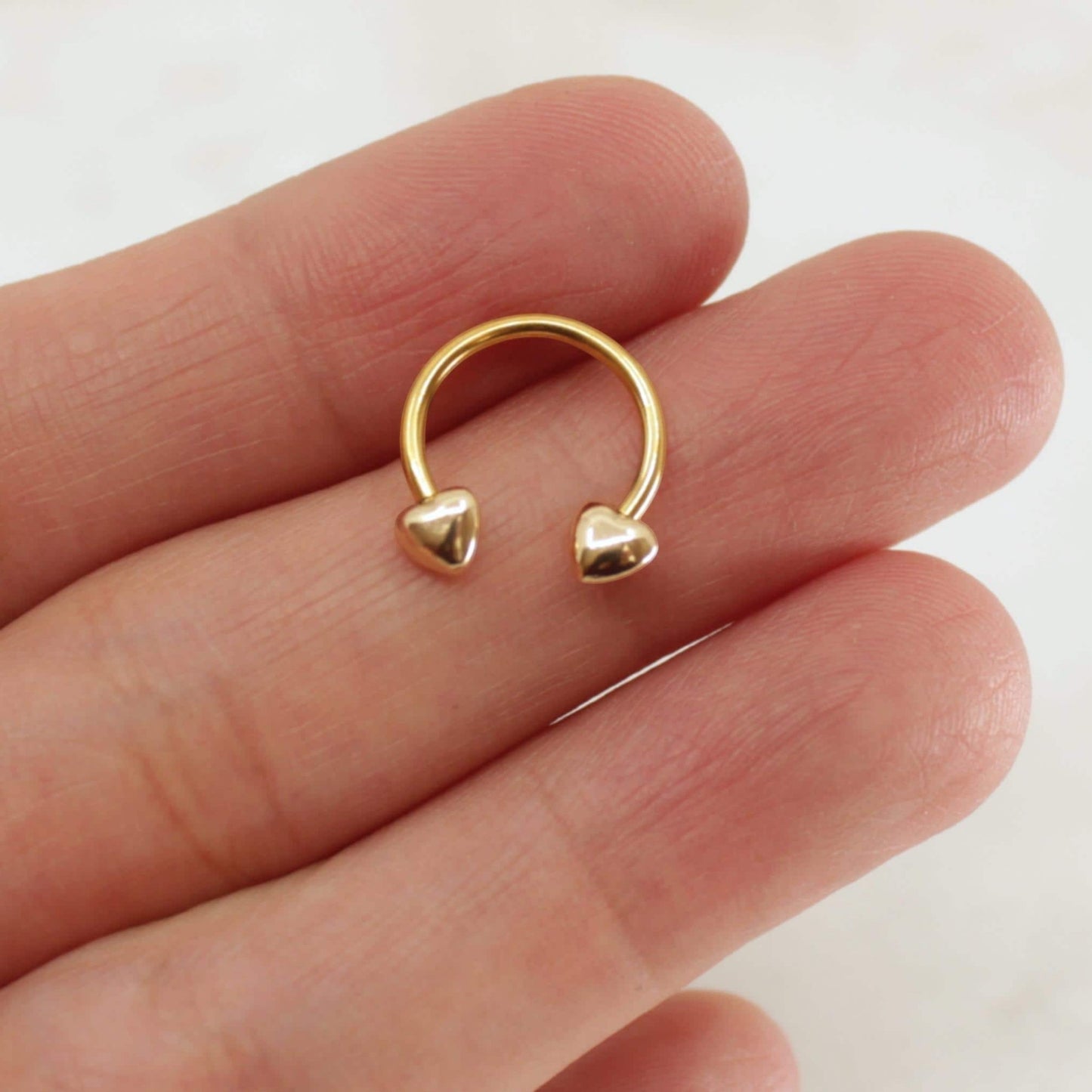 MKBodyJewelry 16G gold surgical steel horseshoe septum ring with minimalist heart ends held between fingers.