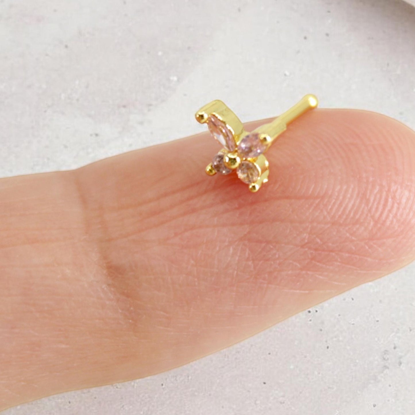 MKBodyJewelry dainty gold nose stud featuring a sparkly pink gemstone butterfly design on a fingertip