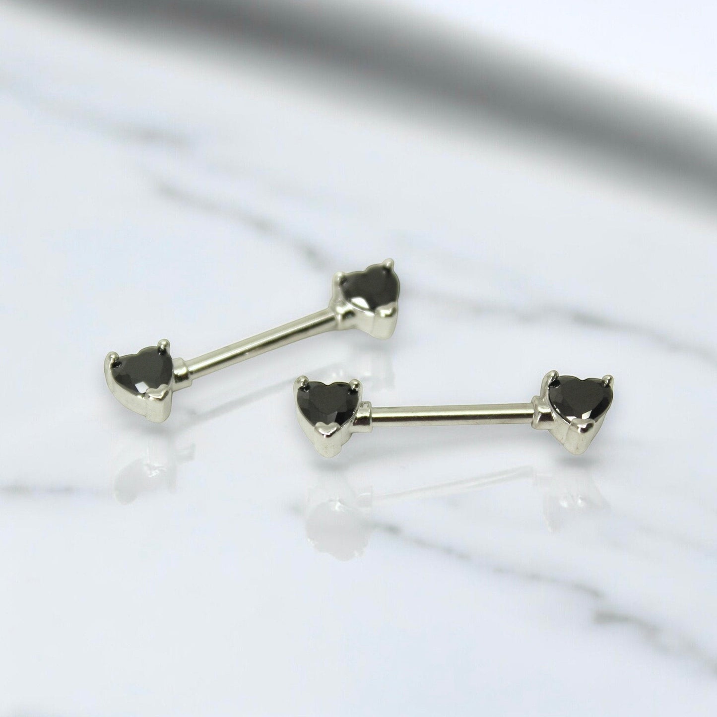 MKBodyJewelry 14g surgical steel nipple barbells featuring black heart-shaped gemstone ends on a white marble surface.