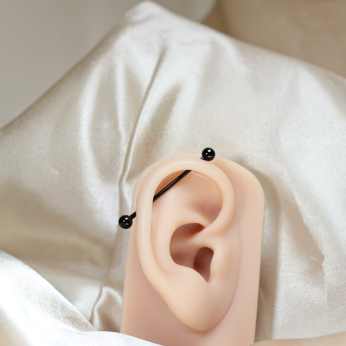 MKBodyJewelry 14G black titanium industrial barbell piercing on a silicone ear model against a satin background.