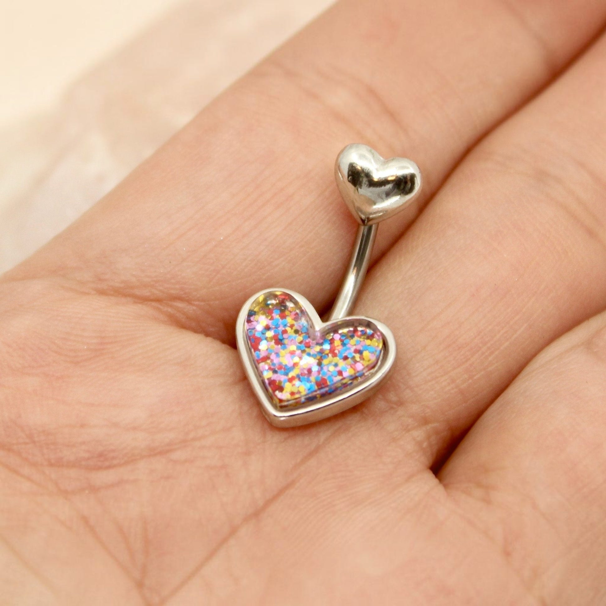 MKBodyJewelry 14G surgical steel belly button ring with a colorful glitter resin heart charm held in a hand.