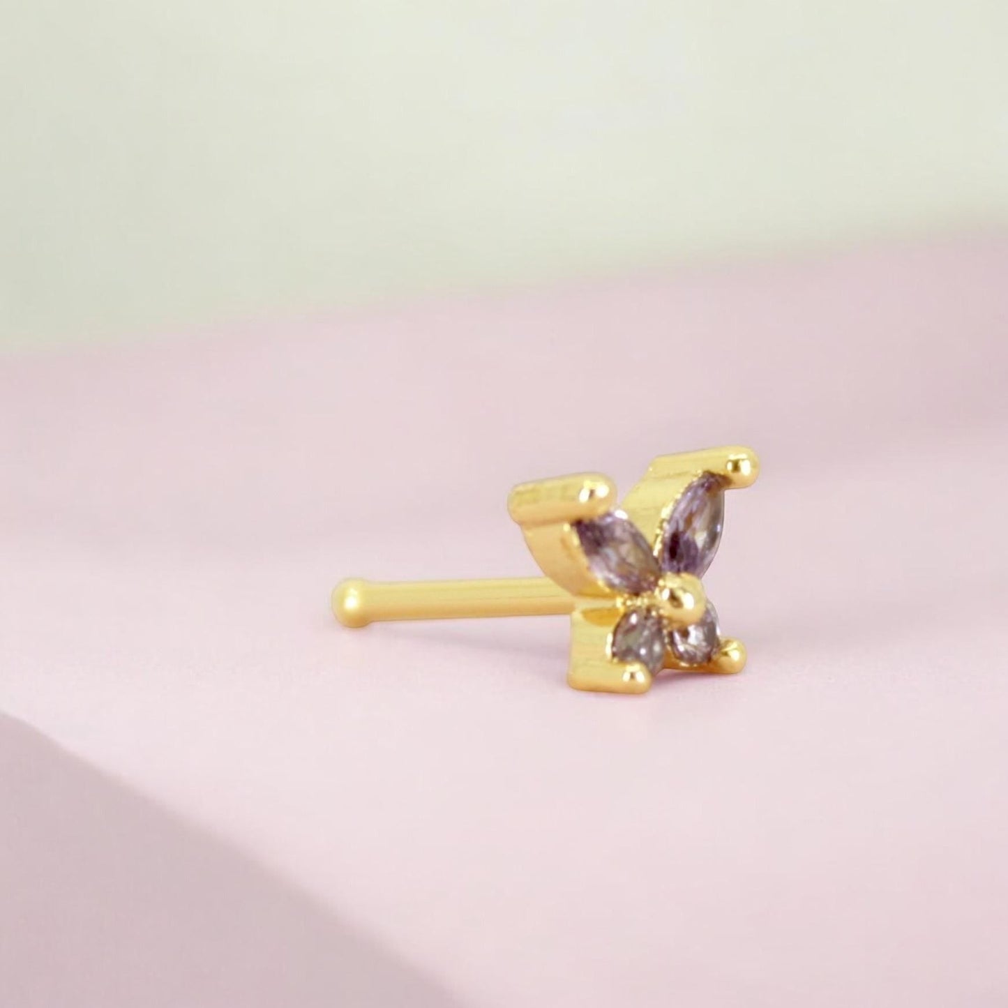 MKBodyJewelry dainty gold nose stud featuring a purple gemstone butterfly design on a light pink background.