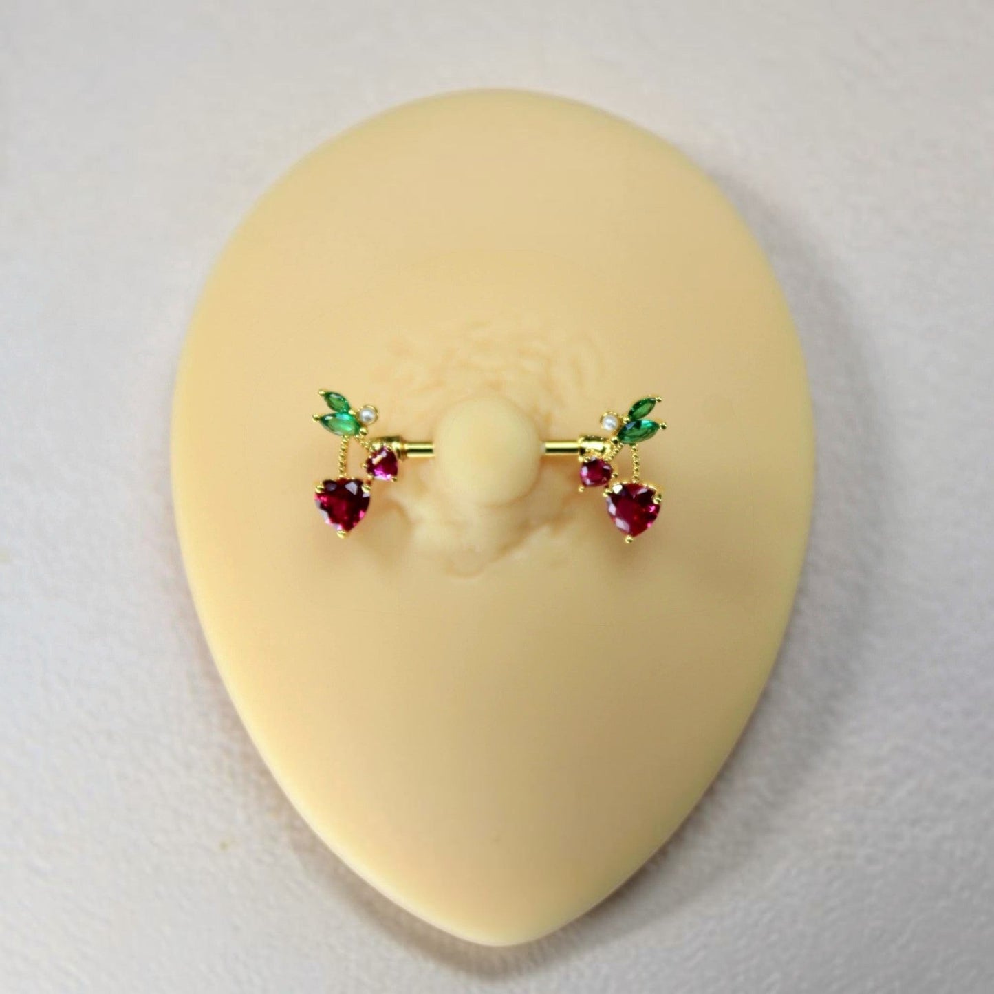 MKBodyJewelry gold cherry heart nipple barbell set with red and green crystal accents on a silicone display model.