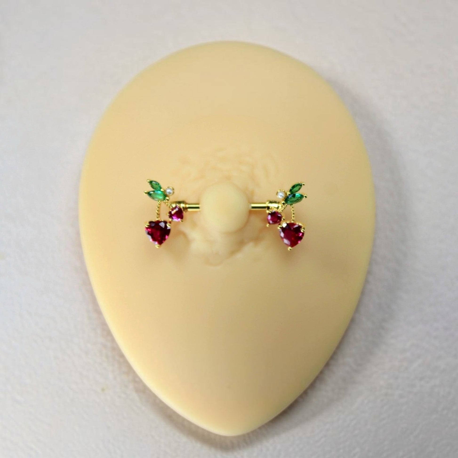 MKBodyJewelry gold cherry heart nipple barbell set with red and green crystal accents on a silicone display model.