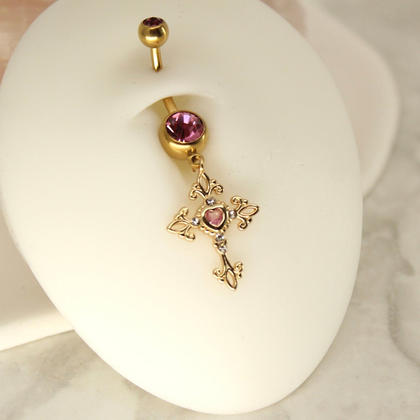 MKBodyJewelry gold tone belly ring featuring a hot pink gem and an ornate dangling cross charm on a surgical steel bar.