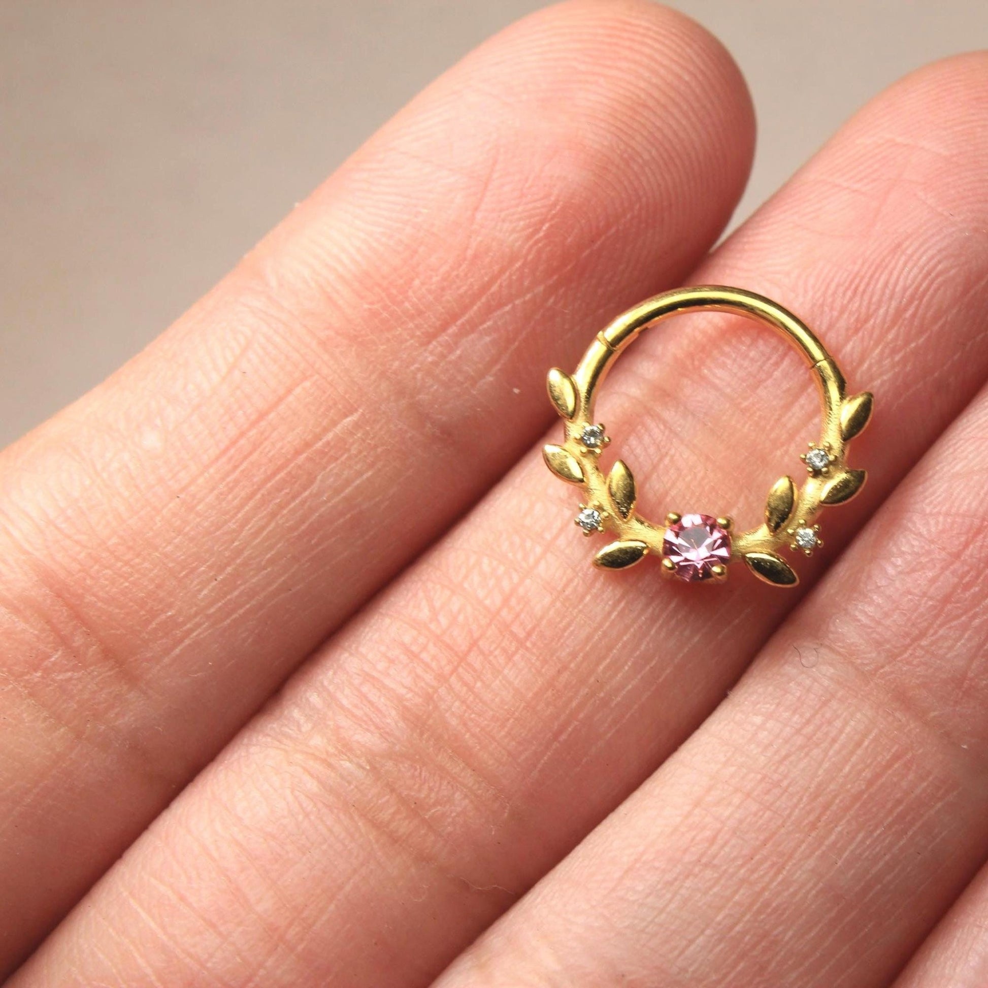 MKBodyJewelry gold floral septum clicker ring with a pink center gem and leaf accents held on a finger.