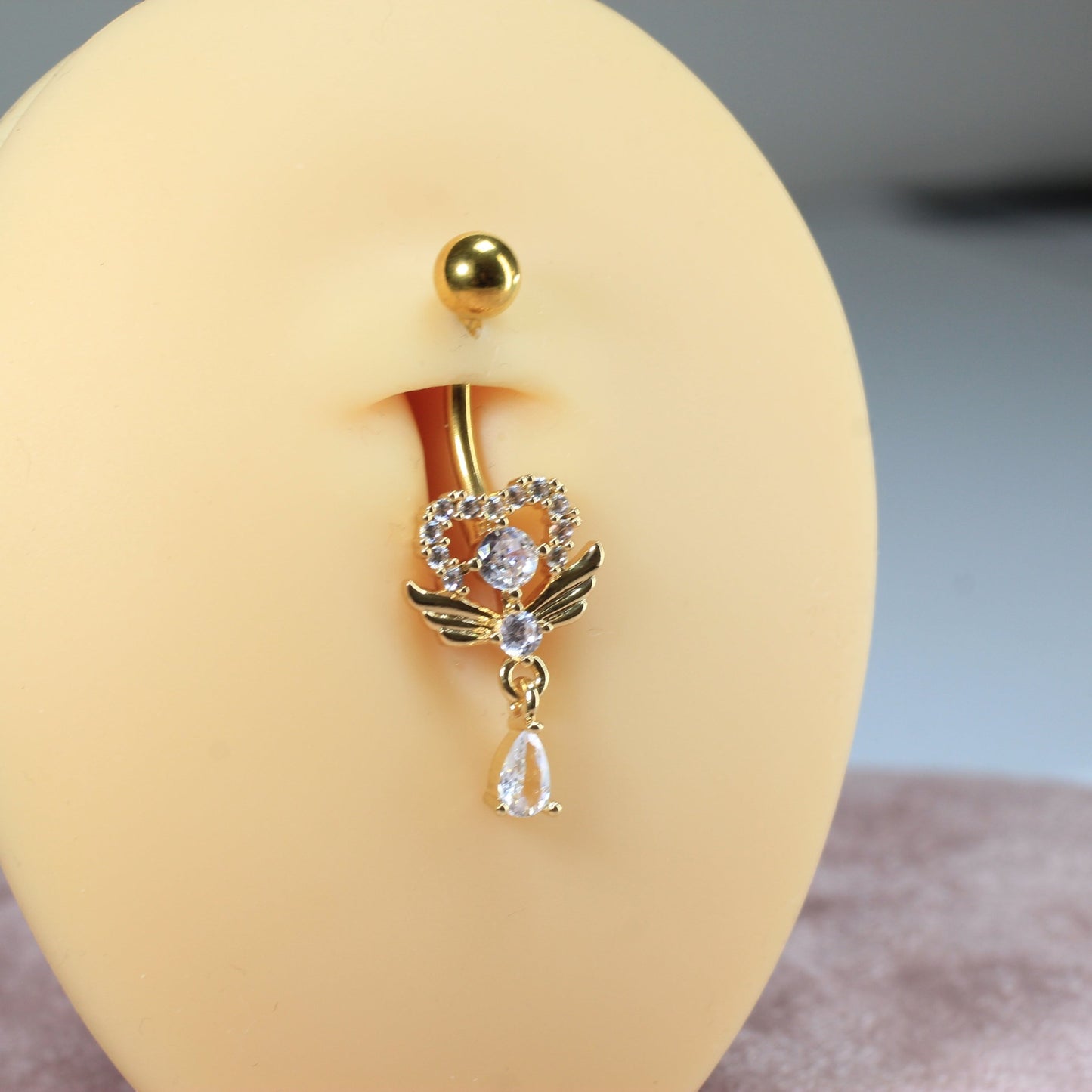 MKBodyJewelry gold heart belly button ring with angel wings and a dangling teardrop crystal on a navel display model.