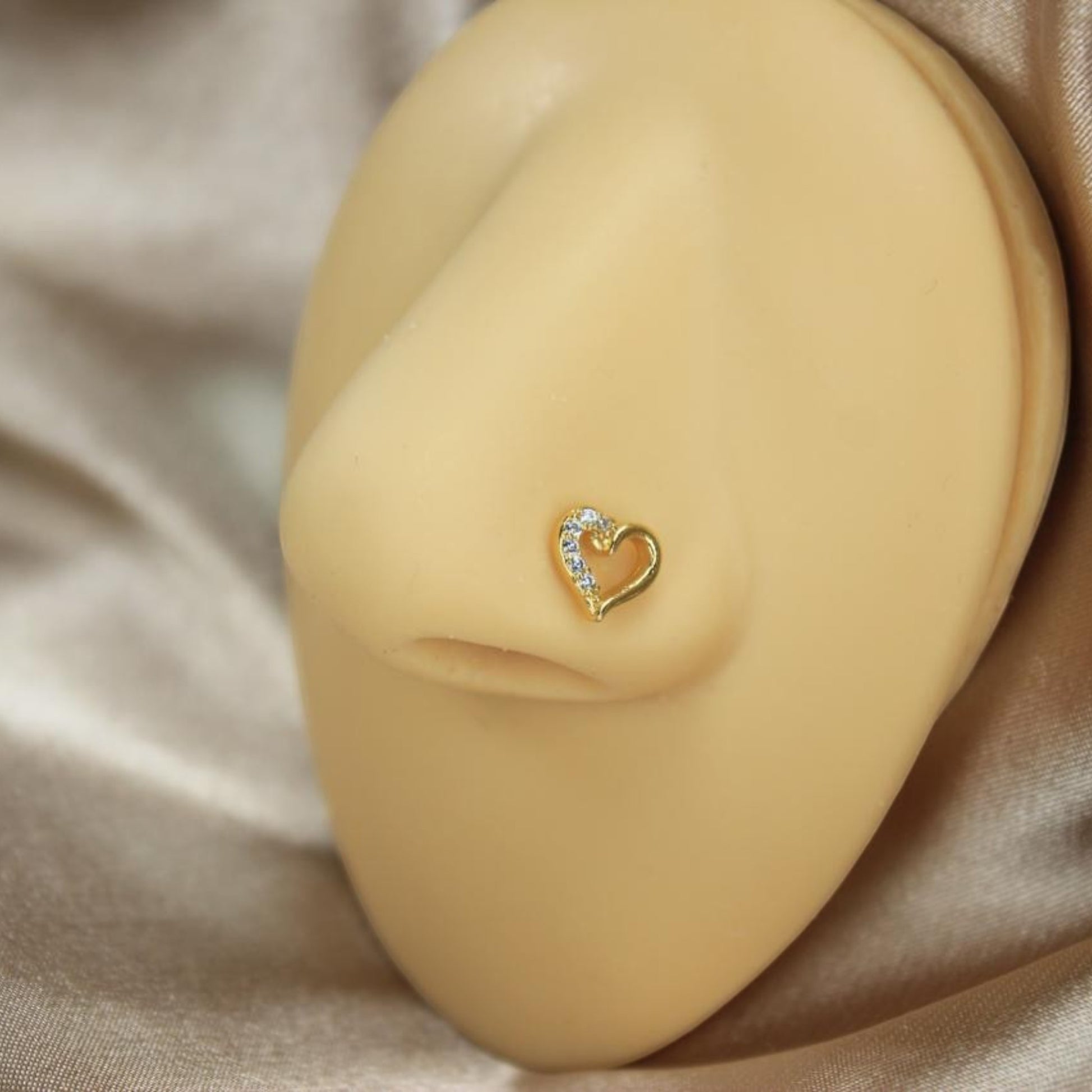MKBodyJewelry 20G gold heart nose stud with sparkling CZ gems on a mannequin nose against a satin background.