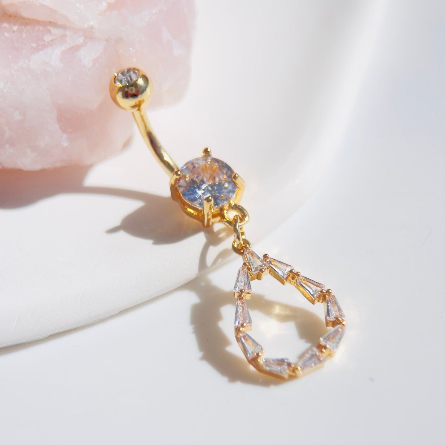 MKBodyJewelry 14G gold dangling belly button ring with a teardrop baguette CZ stone charm on a white surface.