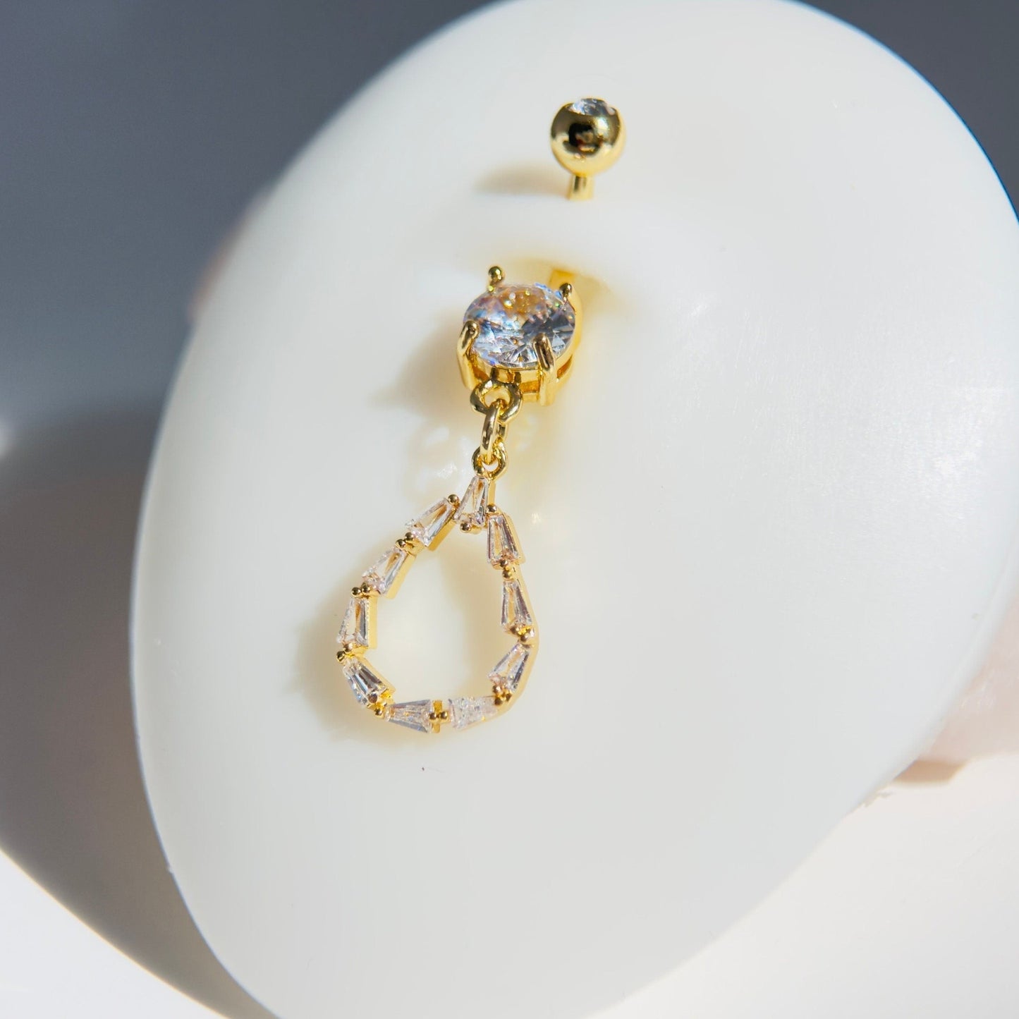MKBodyJewelry 14G gold surgical steel belly button ring featuring a dainty teardrop baguette CZ gem dangle charm.