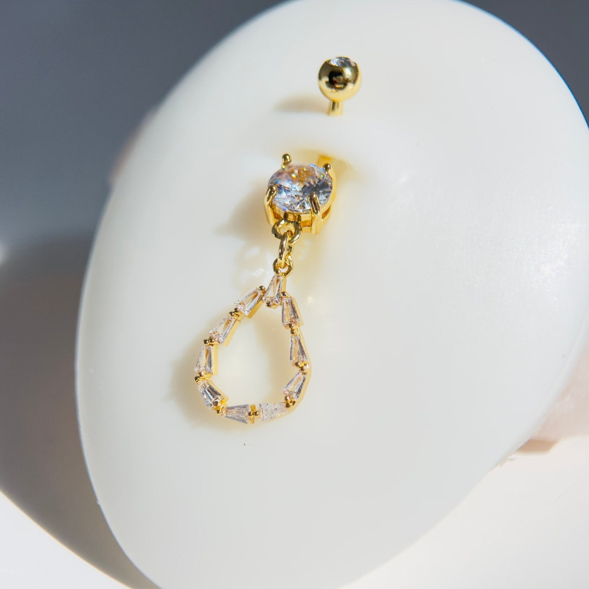 MKBodyJewelry 14G gold surgical steel belly button ring featuring a dainty teardrop baguette CZ gem dangle charm.