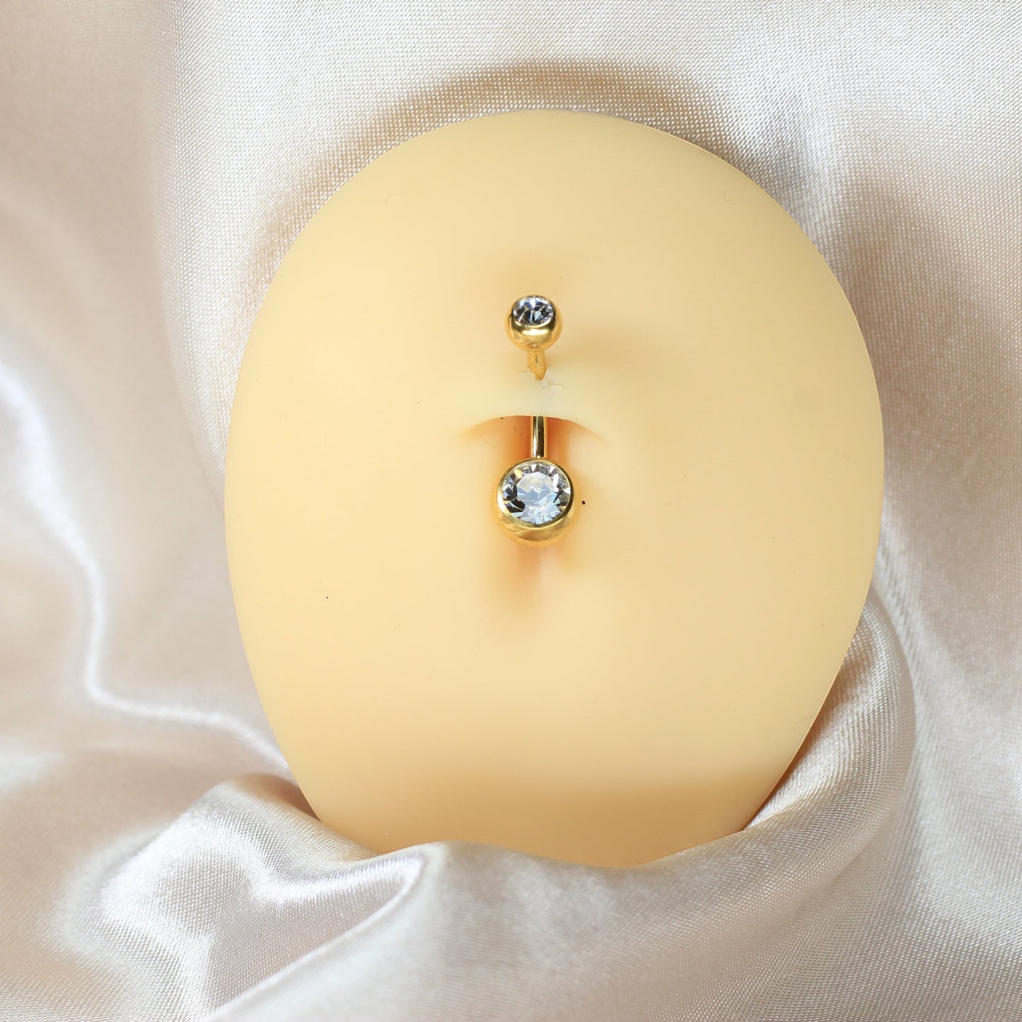 MKBodyJewelry gold titanium belly button ring with double CZ jewels on a realistic navel model against silk fabric.