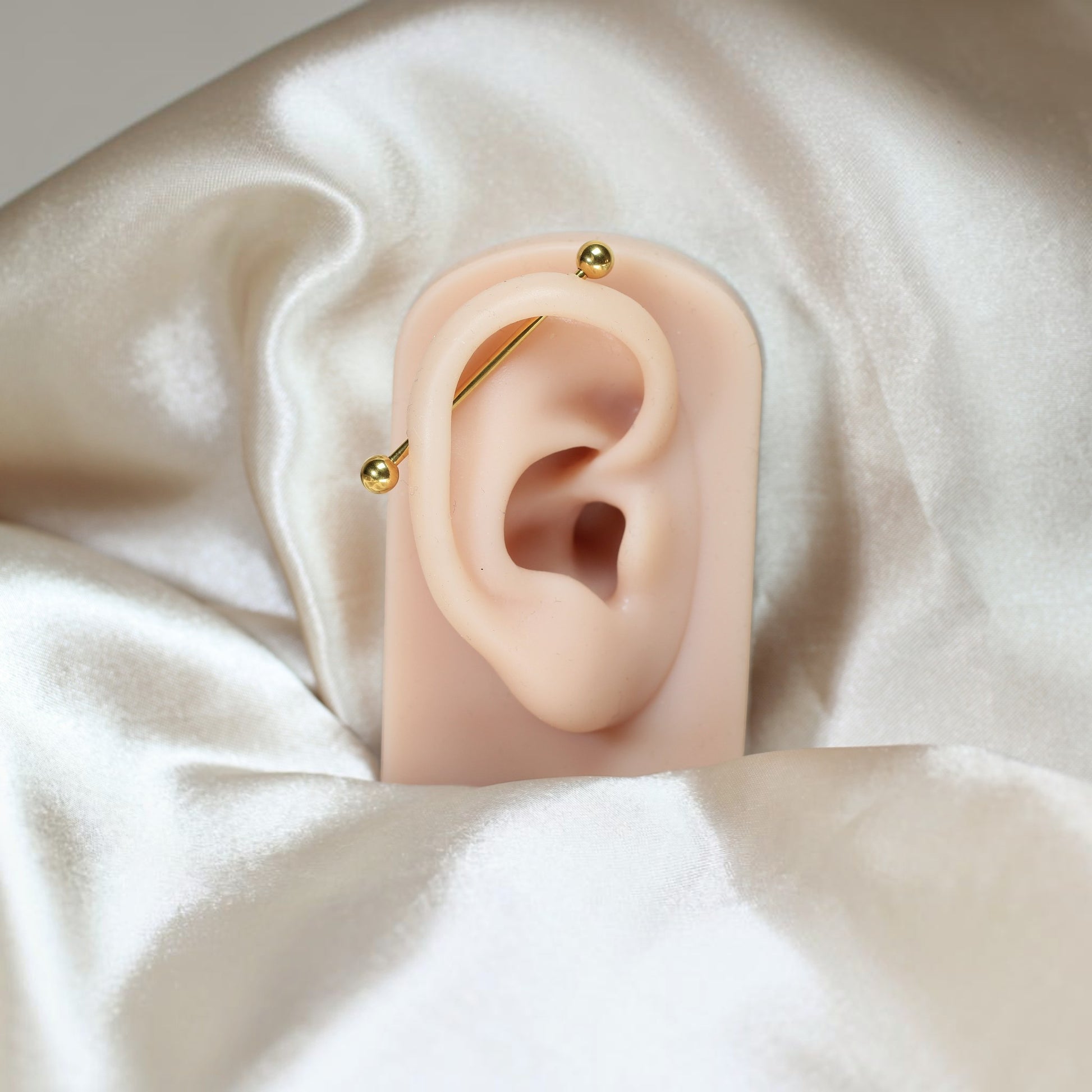 MKBodyJewelry 14G gold titanium industrial barbell piercing on a mannequin ear against a soft satin background.