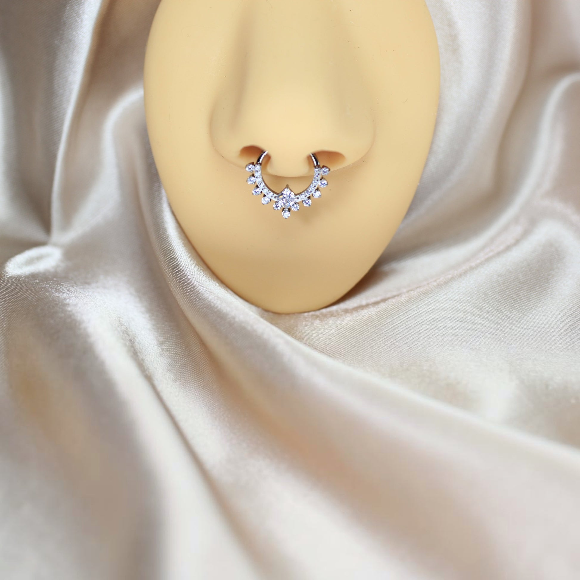 MKBodyJewelry silver titanium septum clicker ring with sparkling CZ stones on a mannequin nose against silk fabric.