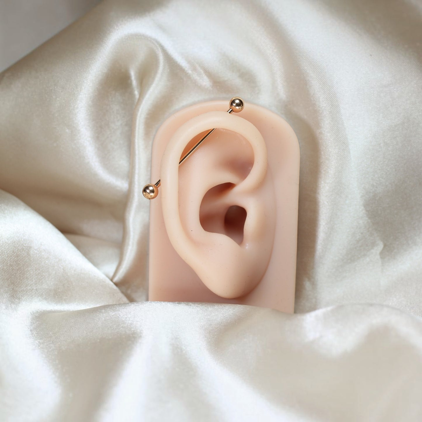 MKBodyJewelry 14G titanium industrial barbell piercing on a mannequin ear, displayed on elegant cream satin fabric.